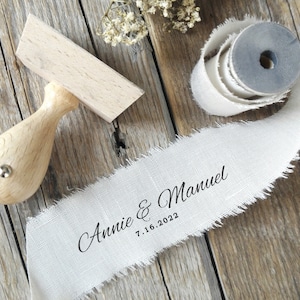 May include: A wooden stamp and a roll of off-white ribbon with frayed edges. The ribbon is printed with the names "Annie & Manuel" and the date "7.16.2022" in black script. The stamp has a wooden handle.