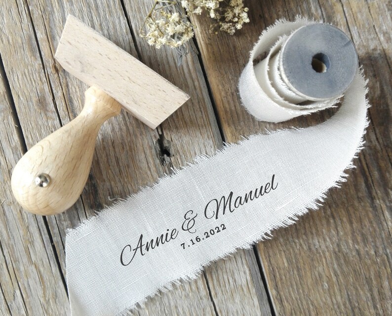 May include: A wooden stamp and a roll of off-white ribbon with frayed edges. The ribbon is printed with the names "Annie & Manuel" and the date "7.16.2022" in black script. The stamp has a wooden handle.