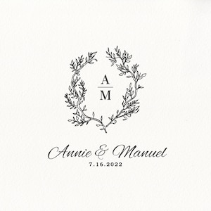 May include: A cream-colored card features a black floral wreath design with the initials "A" and "M" inside. Below the wreath, the names "Annie & Manuel" and the date "7.16.2022" are elegantly scripted. This design is suitable for wedding invitations.