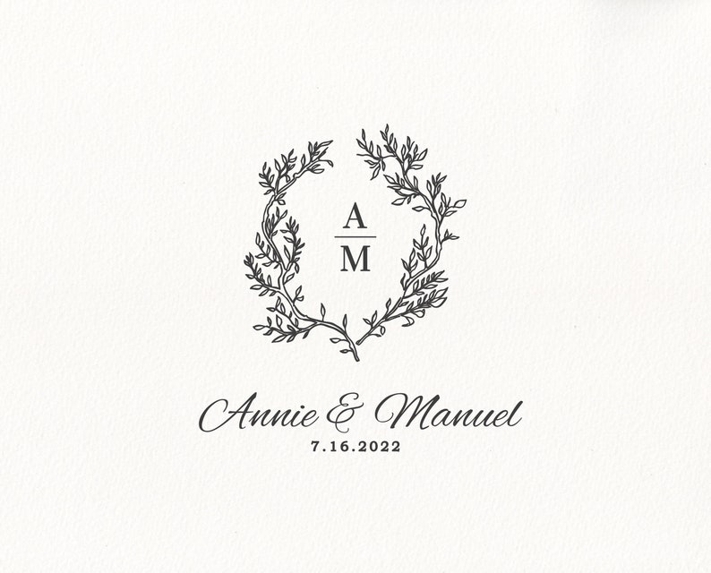 May include: A cream-colored card features a black floral wreath design with the initials "A" and "M" inside. Below the wreath, the names "Annie & Manuel" and the date "7.16.2022" are elegantly scripted. This design is suitable for wedding invitations.