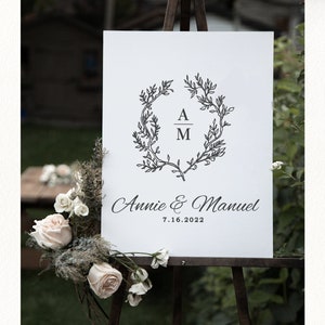 May include: A white wedding welcome sign on an easel. The sign features a black floral wreath with the initials "A" and "M" inside. The names "Annie & Manuel" and the date "7.16.2022" are written below.