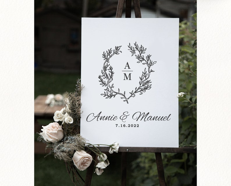 May include: A white wedding welcome sign on an easel. The sign features a black floral wreath with the initials "A" and "M" inside. The names "Annie & Manuel" and the date "7.16.2022" are written below.