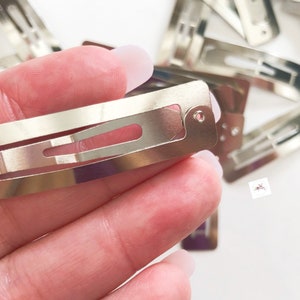 May include: Silver-colored metal snap hair clips. The rectangular clips have a simple design with a slotted opening and a small hole at one end. Several clips are visible, some in focus, others blurred in the background.