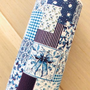 May include: Rolled-up fabric with a patchwork design in shades of blue and white. The pattern includes floral prints, gingham, stripes, and a central square with a blue bow and leaves. The fabric is likely for crafts or home decor.