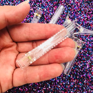 May include: A close-up of a clear plastic hair clip with a metal spring. The clip is held in a hand against a sparkly background.