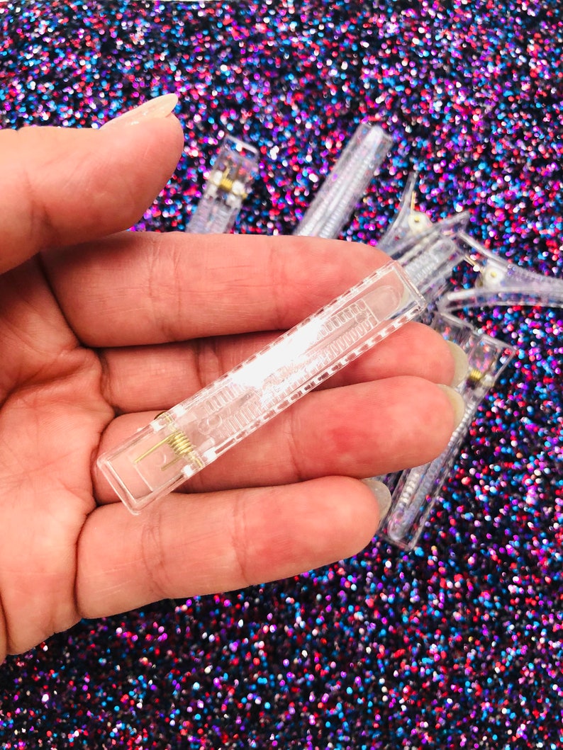 May include: A close-up of a clear plastic hair clip with a metal spring. The clip is held in a hand against a sparkly background.