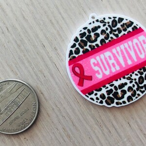 May include: A round pink and white sticker with a leopard print background. A pink ribbon with the word "SURVIVOR" in white letters is on the sticker.