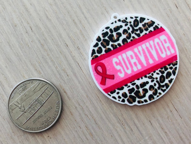 May include: A round pink and white sticker with a leopard print background. A pink ribbon with the word "SURVIVOR" in white letters is on the sticker.