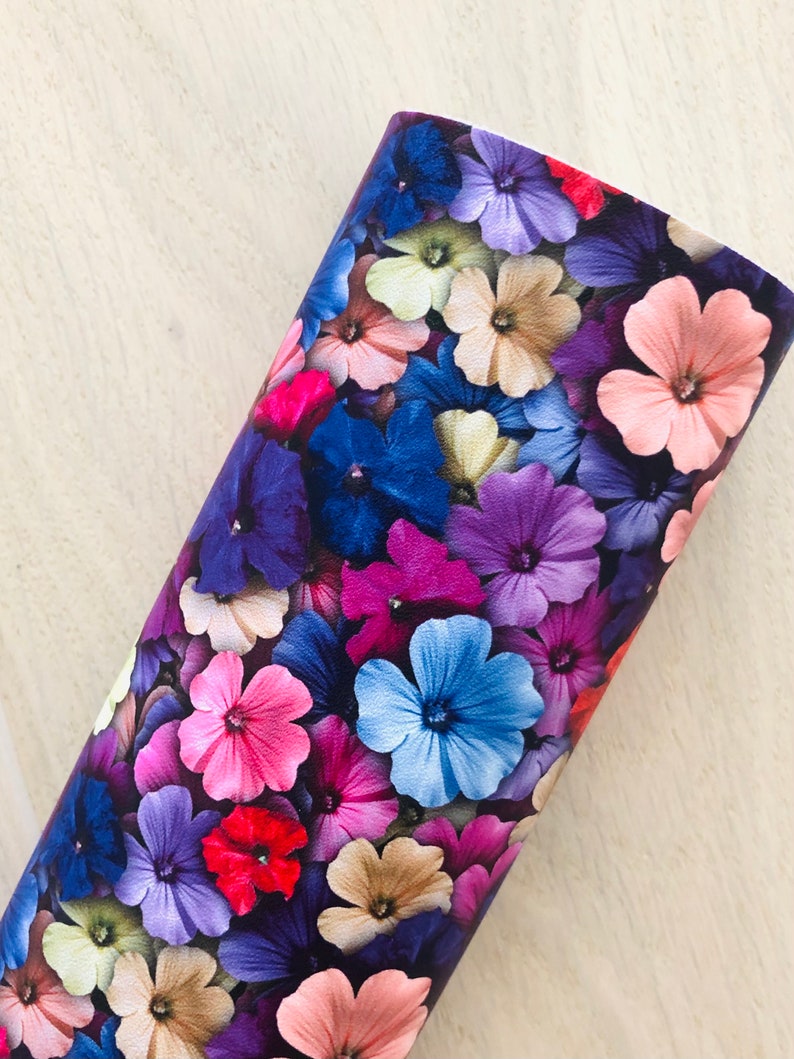 May include: A close-up of a fabric with a floral print. The fabric features a variety of flowers in shades of pink, purple, blue, and beige. The flowers are arranged in a random pattern and appear to be overlapping.
