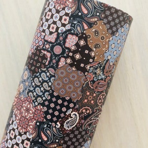 May include: A rolled-up fabric with a patchwork design. The pattern features various shapes and colors, including paisley, floral, and geometric motifs in shades of brown, black, blue, and pink. The fabric appears to be leather or a similar material.