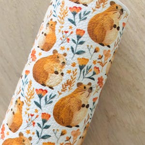 May include: A roll of white faux leather featuring a repeating pattern of brown hamsters, orange flowers, and green leaves. The design is printed on a textured surface, creating a whimsical and nature-inspired aesthetic. The roll is displayed on a light wood surface.