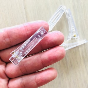 May include: A close-up of a clear plastic hair clip. The clip is held in a hand. The clip has a metal spring.