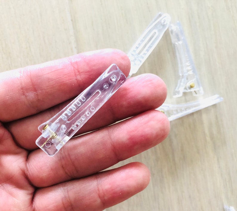 May include: A close-up of a clear plastic hair clip. The clip is held in a hand. The clip has a metal spring.
