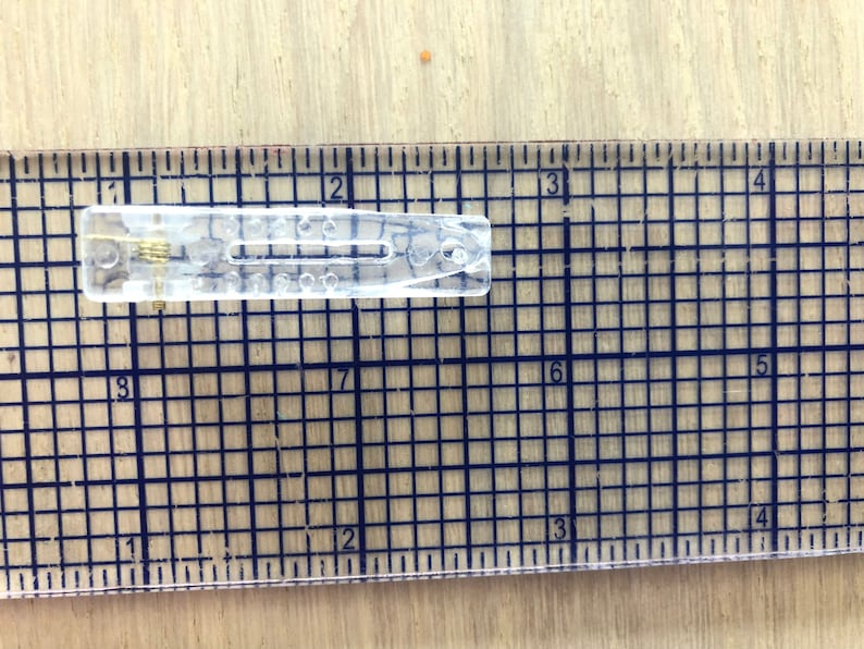 May include: A clear plastic hair clip with a metal spring. The clip is about 2 inches long and is measured against a ruler.