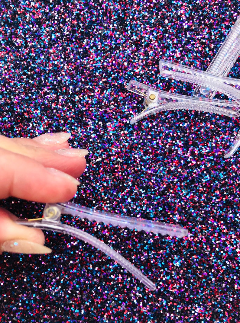 May include: Clear plastic hair clips on a sparkly blue, red, and black background. The clips have a gold metal spring.