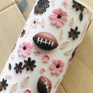 May include: White fabric with a brown and pink floral pattern and brown footballs. The fabric has a textured surface.