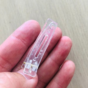 May include: A clear plastic hair clip with a metal spring. The clip is designed to hold hair in place.