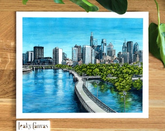 May include: A watercolor painting of the Philadelphia skyline with a view of the Schuylkill River and a walking path. The city buildings are in the background and the river is in the foreground. The painting is done in shades of blue, green, and brown.