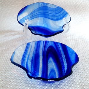 May include: Two shallow glass bowls featuring a vibrant blue and white swirled pattern. The bowls have an organic, wavy rim shape and a smooth, glossy finish. One bowl is positioned upright on a clear stand, while the other sits below it.