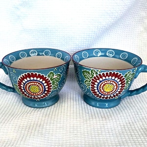 May include: Two matching ceramic mugs with a light blue exterior and a cream-colored interior. Each mug features a large, hand-painted floral design in red, yellow, and white, accented by green leaves and white swirl patterns. The mugs have a pedestal base.