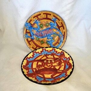 May include: Two round ceramic plates featuring lizard motifs. The top plate displays a blue lizard on a yellow and red geometric background. The bottom plate features two red lizards on a yellow background with blue and red geometric patterns.
