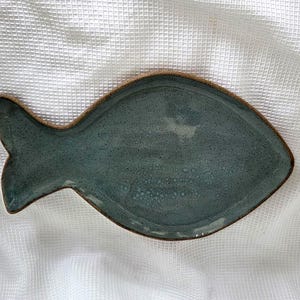 May include: A fish-shaped ceramic dish with a glossy, speckled blue-grey glaze and a visible brown clay rim. The surface of the dish features a smooth, slightly uneven texture with subtle variations in color depth across the body and tail.