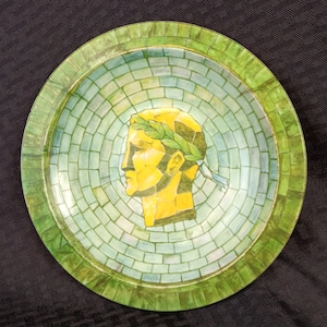 May include: A round, green and blue ceramic plate with a mosaic design. The center of the plate features a yellow, painted profile of a person wearing a laurel wreath.