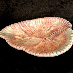 May include: A ceramic leaf-shaped dish with a textured surface. The dish features a color palette of peach, pink, and green. The edges are scalloped, and the dish has a central vein pattern.