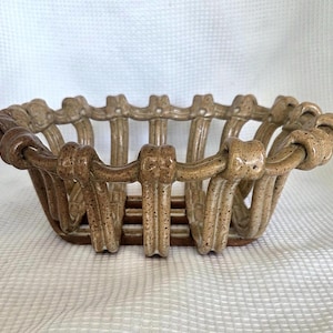May include: A ceramic basket with an openwork design featuring vertical ribbed slats and a looped rim. The piece has a speckled tan and brown glaze finish, with a structured, rectangular base and curved sides that create a woven appearance.