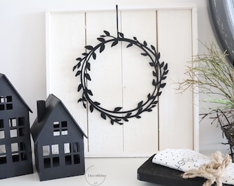 May include: Black and white wreath with a simple leaf design hanging on a white wooden wall. Two black houses are in the foreground.