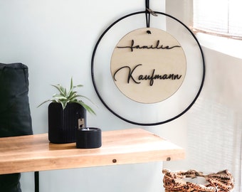 Puede incluir: Un letrero redondo de madera con el texto "Familie Kaufmann" en letras cursivas negras, colgado de un aro de metal negro. El letrero está en una pared blanca con un estante de madera debajo.