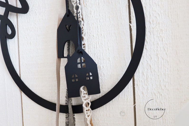 Puede incluir: Adornos de madera negra en forma de casa con ventanas recortadas que cuelgan de un aro de metal negro con cuerda blanca y marr&oacute;n. El texto "Decofieber" es visible en la esquina inferior derecha.