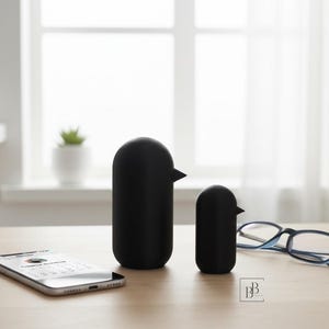 Könnte beinhalten: Zwei schwarze, vogelartige Dekorationsgegenstände, einer groß und einer klein, stehen auf einer Holzoberfläche. Ein Smartphone und eine Brille liegen ebenfalls auf dem Tisch. Die Objekte haben ein minimalistisches Design mit einem schnabelartigen Vorsprung.