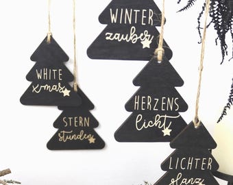 Puede incluir: Cinco adornos navideños de madera negra que cuelgan de cordeles de yute. Cada adorno presenta texto en blanco y un pequeño gráfico de estrella. El texto dice: WHITE xmas, STERN stunden, WINTER zauber, HERZENS licht y LICHTER glanz.