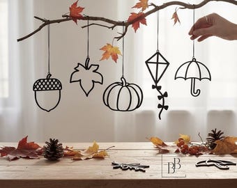 May include: Five black hanging ornaments shaped like an acorn, a maple leaf, a pumpkin, a kite, and an umbrella, suspended from a branch with autumn leaves. Two additional flat black shapes lie on a wooden surface with pinecones and foliage.