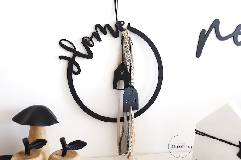 Puede incluir: Decoraci&oacute;n para el hogar de metal negro con la palabra "home" en una fuente de escritura, un c&iacute;rculo y dos peque&ntilde;os adornos negros en forma de casa colgando de &eacute;l. El c&iacute;rculo est&aacute; colgado en una pared blanca.