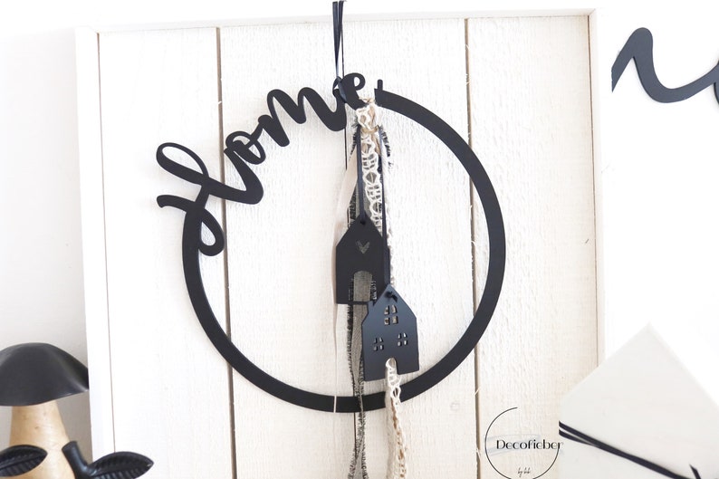 Puede incluir: Decoraci&oacute;n para el hogar de metal negro con una corona y la palabra "home" en una fuente de escritura. La corona tiene dos adornos negros en forma de casa colgando de ella.