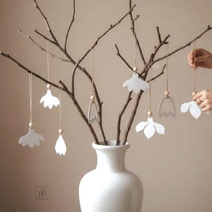 Könnte beinhalten: Eine weiße Keramikvase hält Zweige, die mit hängenden weißen Blumenschmuck verziert sind. Die Ornamente sind an dünnen Schnüren mit Holzperlen aufgehängt. Eine Hand justiert eines der Ornamente. Der Hintergrund ist ein neutrales Beige.