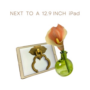 Puede incluir: Una flor de lirio blanco y rosa en un jarrón de vidrio verde junto a un iPad de 12,9 pulgadas con un aldaba de puerta dorada en la pantalla.