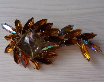 Peut inclure: Une broche dorée avec un grand cristal iridescent au centre, entouré de petits cristaux irisés dans un motif en forme de feuille.