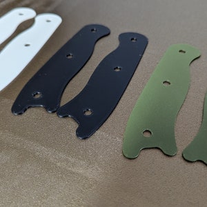 May include: Three pairs of knife scales, one white, one black, and one green. The scales are made of a smooth material and have a curved shape.