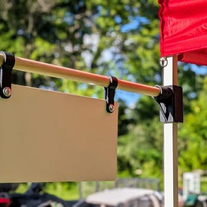 May include: A rectangular blank sign panel suspended from a horizontal wooden rod. The sign is attached to the rod with two black clips and metal hardware. The rod is mounted to a vertical support pole, positioned beneath a bright red canopy fabric.