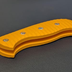 May include: A bright yellow knife handle with a grooved texture and silver rivets. The handle is made of two curved pieces, creating a comfortable grip. The knife handle is shown against a dark background.