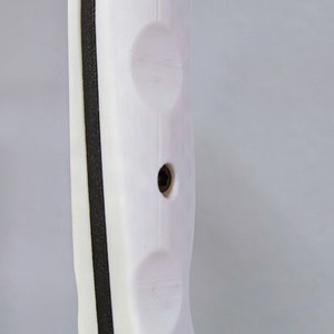 May include: White knife handle with black accents and four screw holes. The handle is made of a textured material and has a rounded shape. The text "K-BAR USA" is visible on the bottom of the handle.