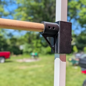 May include: Close-up of a wooden dowel rod held in place by a black metal bracket attached to a white support post. The background includes a red truck and green foliage.