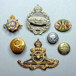 Op de afbeelding: Verzameling vintage militaire insignes en knopen in goud, zilver en email. Inclusief een tankembleem, een kanonembleem en diverse knopen met kroon- en vogelmotieven. Voor verzamelaars.
