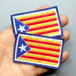 May include: Two rectangular patches with a flag design. The flag has horizontal red and yellow stripes, a blue triangle with a white star. The patches have a blue border and a white stitched edge. The patches are held in a hand.