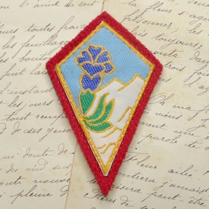 May include: A red, white, and blue embroidered patch with a stylized flower design. The patch is diamond-shaped and has a white background with a blue flower and green leaves.