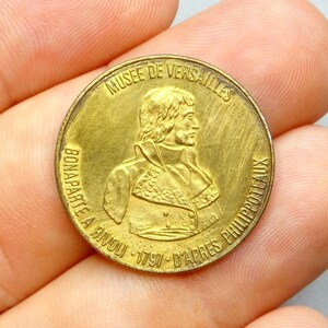 Peut inclure: Une pièce de monnaie dorée avec le profil d'un homme et du texte en français. La pièce porte l'inscription "MUSEE DE VERSAILLES BONAPARTE A RIVOLI 1797 D'APRES PHILIPPOTEAUX". La pièce est tenue dans une main.