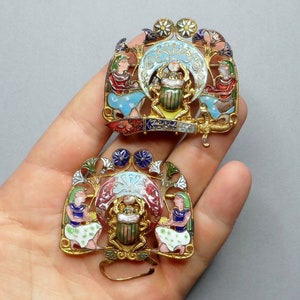 May include: Two gold-toned, ornate, enamel-painted brooches with a scarab beetle design. Each brooch features a crescent moon, stylized flowers, and figures in colorful clothing.